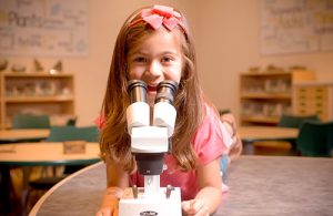 Girl with pink shirt and bow on her head looking up from a white microscope
