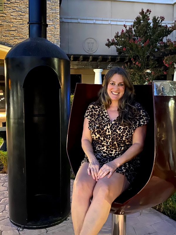 woman sitting in giant, plastic wine glass with giant wine bottle nearby
