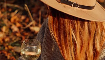 Woman holding a glass of white wine wearing a tan hat with a black belt around it