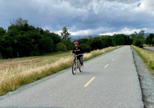 Woman riding a bike on paved path with green grass and trees and clouds