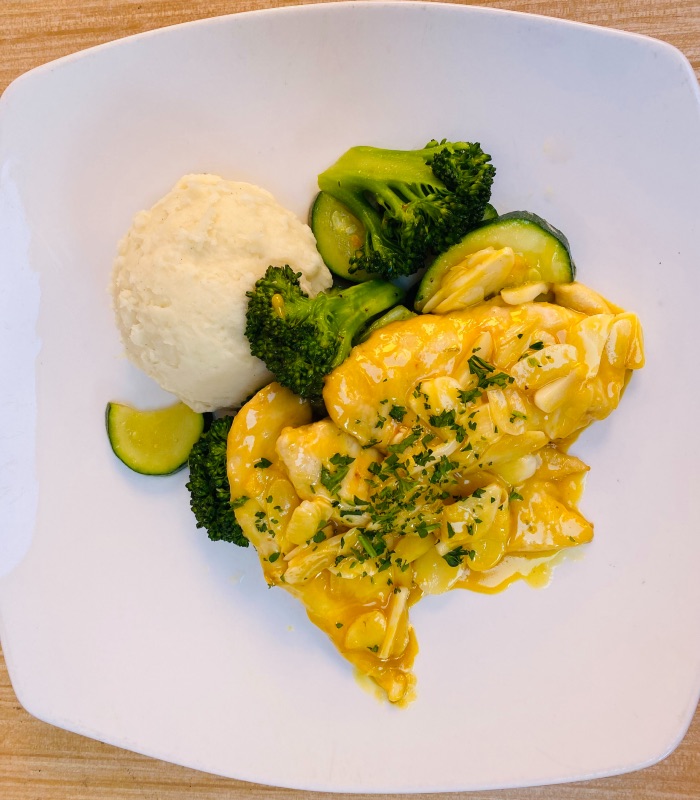 a white plate with mashed potatoes, broccoli, and chicken breasts with garlic cloves on top and a citrus sauce over it