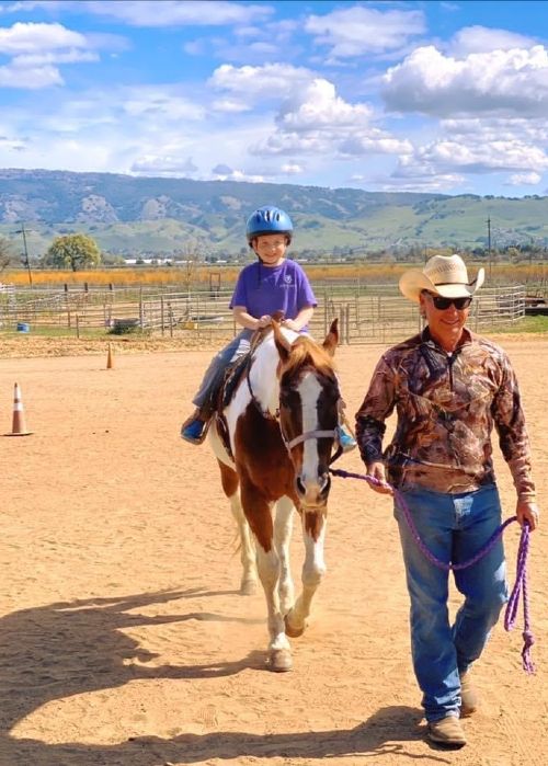 man in cowboy hat leading a horse with a boy in purple shirt on it with green hills in background