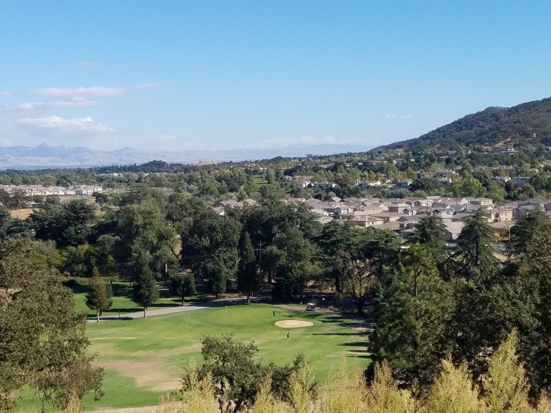 view of a golf course hole surrounded by green grass, tees, and a group of homes in the background against the hills