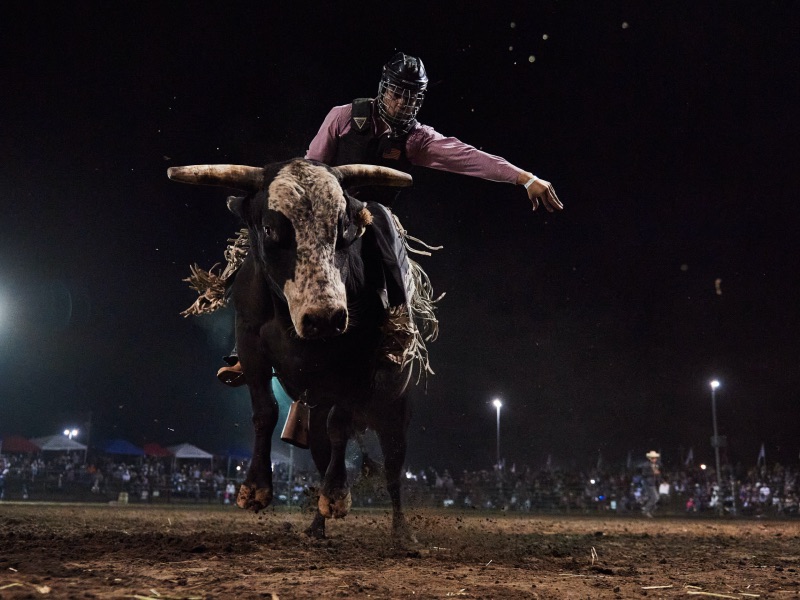 man with helmet riding a brown and white bucking bull with horns with the night sky behind him