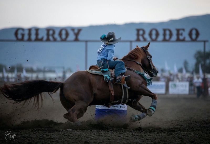 a cowboy riding a brown horse with the Gilroy Rodeo sign in the background