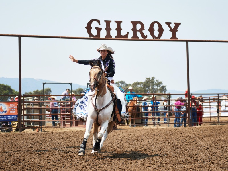 A young lady in a white cowboy horse waving rides an shite and brown horse with the Gilroy rodeo sign behind her