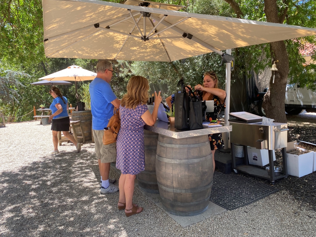 a woman is purchasing wine at an outside bar with woman helping her and two servers in blue shirts are working