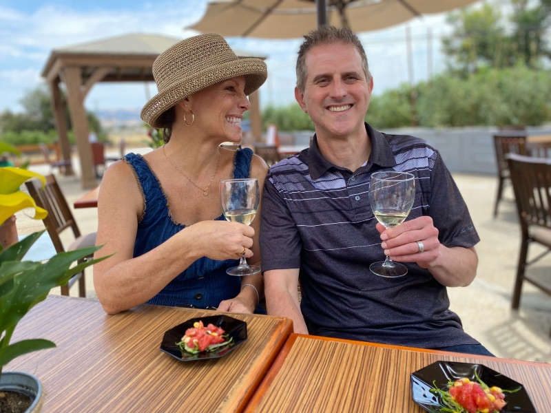 a woman and man smiling while holding glasses of white wine sitting outdoors at a table