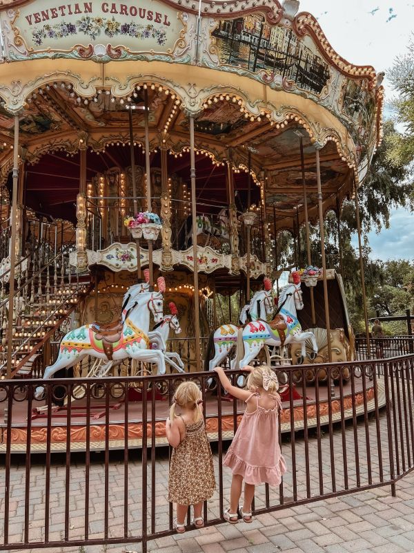 two little blond girls by the fence in front of a two-level carousel