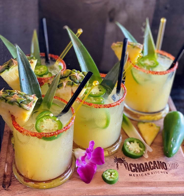 four glasses filled with a yellow-green drink with pineapple and jalapeño garnishes