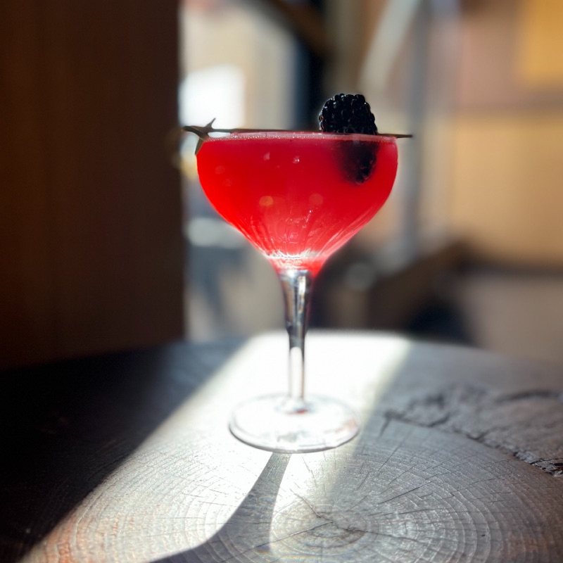 a cocktail glass on a wooden table filled with a red-colored drink with a blackberry garnish on the top