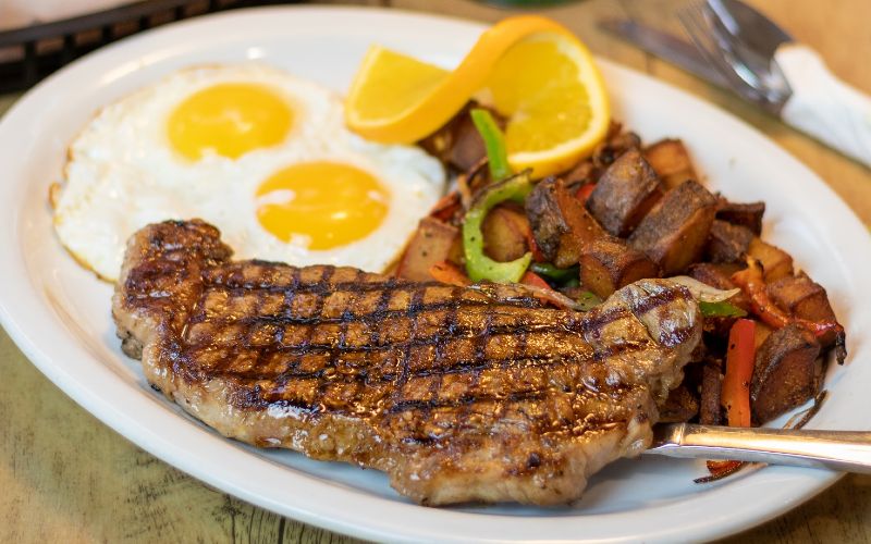 a white plate with a large steak, fried eggs, and potatoes