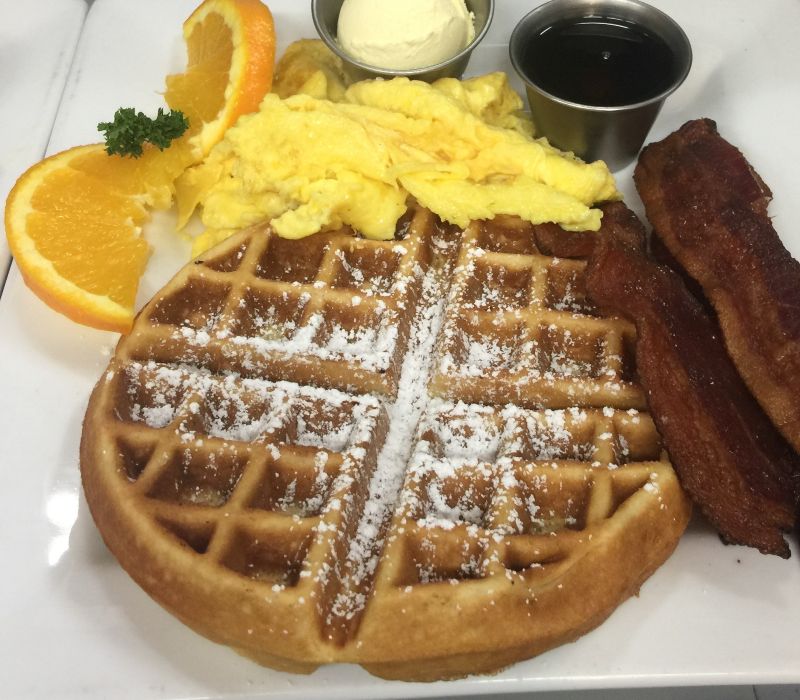 a white plate with a waffle with powdered sugar, scrambled eggs, and bacon
