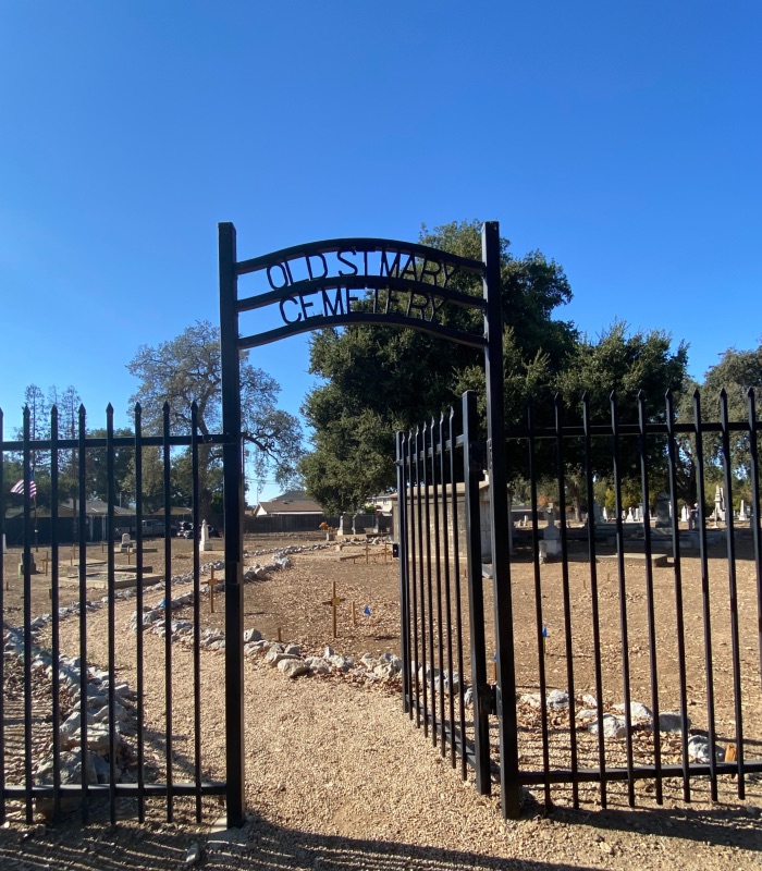 a black iron fence and gate with a dirt path lined by rocks into a cemetery