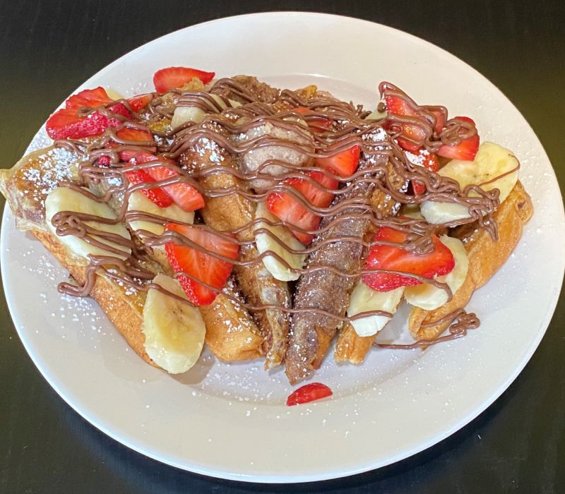 a white plate with French toast with bananas, strawberries, and chocolate sauce drizzled over it all