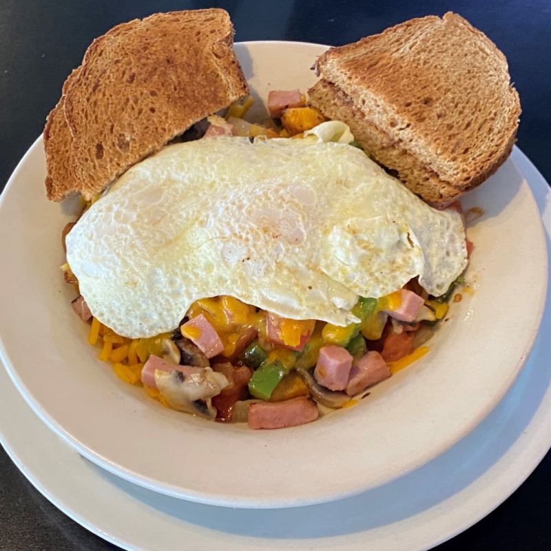a deep white bowl with small bits of meat mixed with veggies, onions, and peppers, topped with cheese and fried eggs and slices of toast