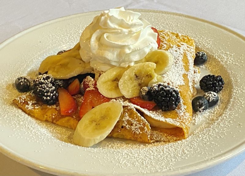 a white plate piled with crepes with berries and bananas and whipped cream