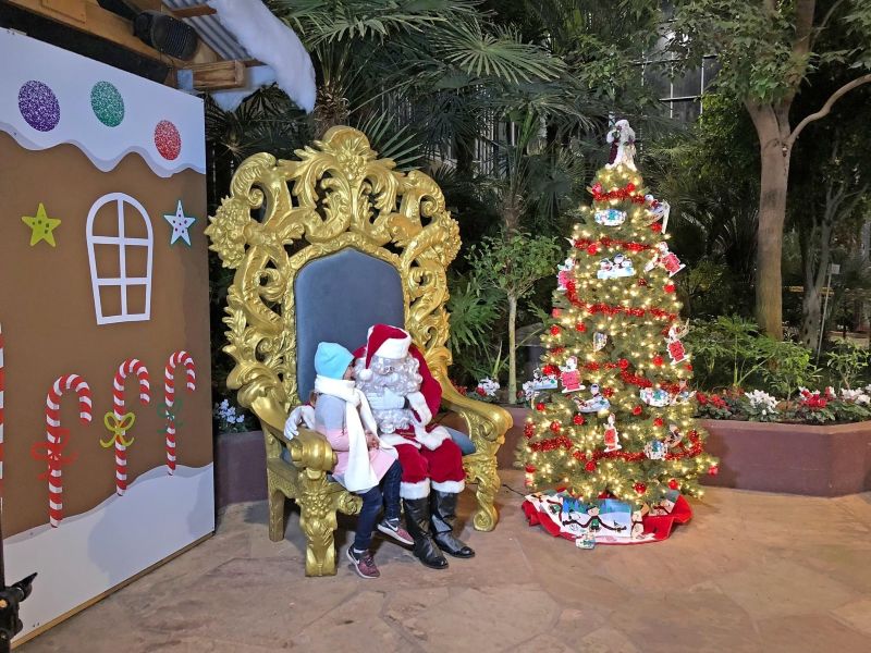 a little girl sitting with Santa on a gold throne with a Christmas tree and a gingerbread house backdrop to the left