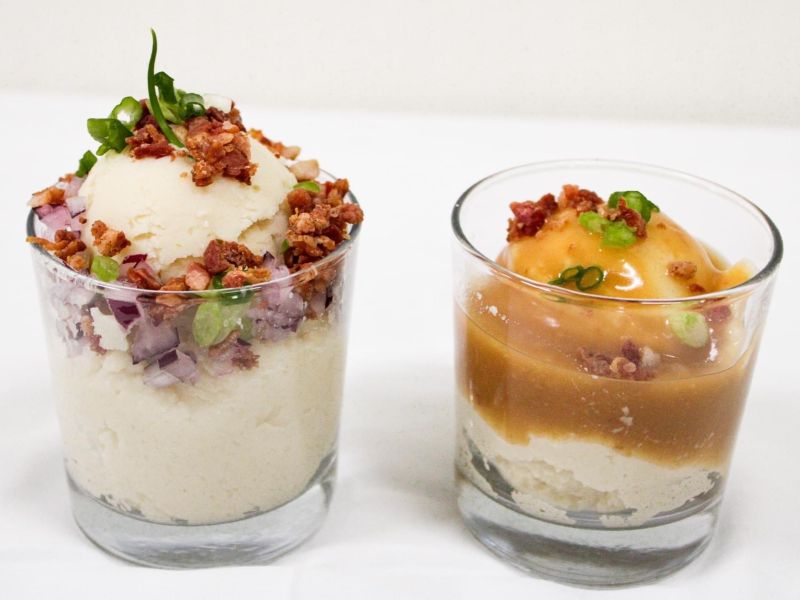 two small glasses with mashed potatoes in them with toppings of gravy, bacon, and onions