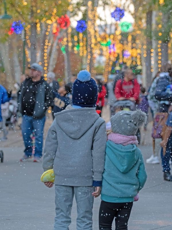 Two children and walking in the fake snow with other people and holiday lights in the background