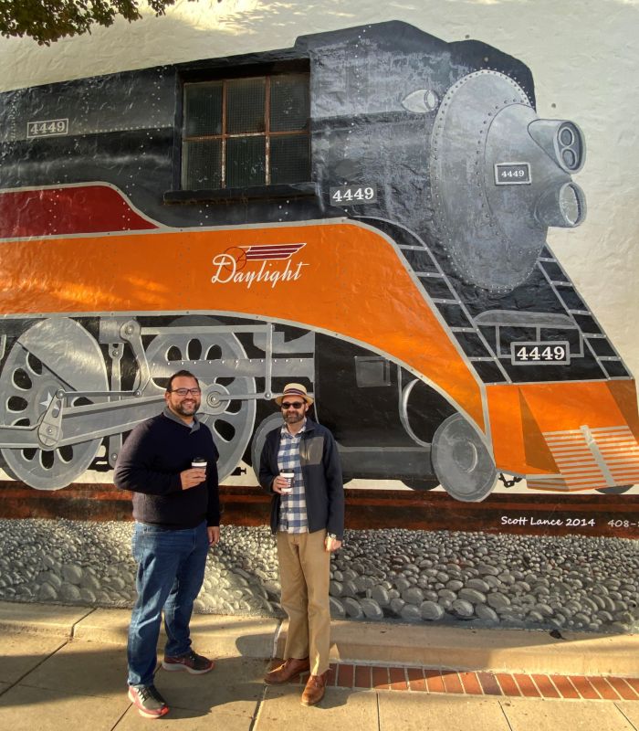 two men holding coffee cups standing in front a train mural