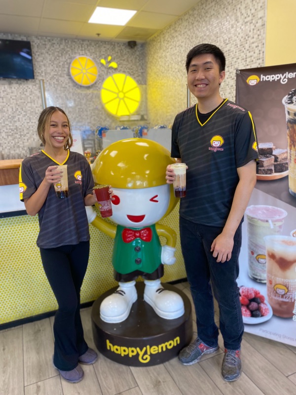 a young woman and a young man pose holding tea drinks with a lemon character smiling in between them