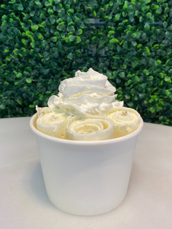 a white cup of rolled white garlic ice cream with whipped cream on top on a white table with green shrub in background