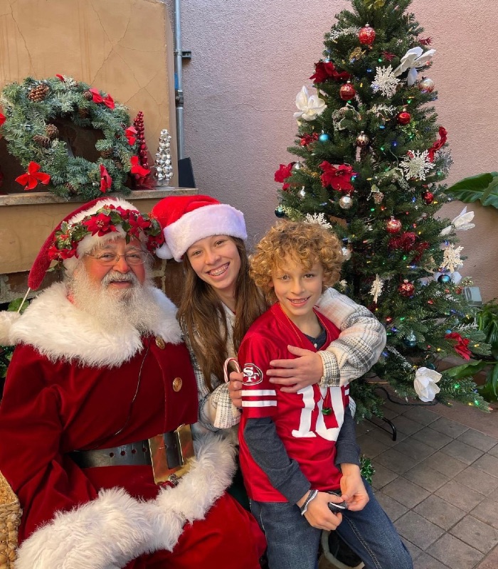a man dressed as Santa in red and white and a young lady and a boy with him with a Christmas tree decorated in the background