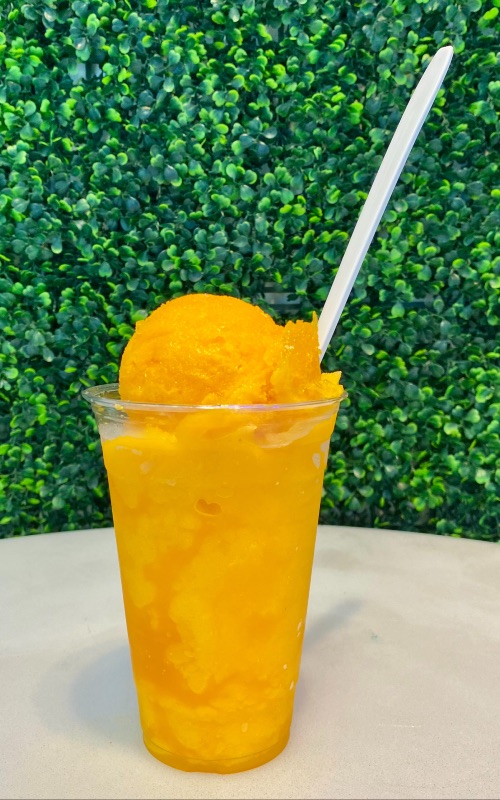 an orange-colored passion fruit slushy in a clear cup with a green shrub background
