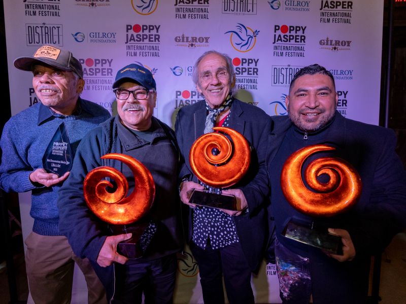 Four men holding orange swirl-shaped awards in front of a Poppy Jasper Film Festival labeled backdrop