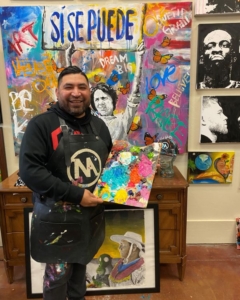 a man smiling in his art studio with a black apron holding a palate with colorful paint on it with paintings in the background