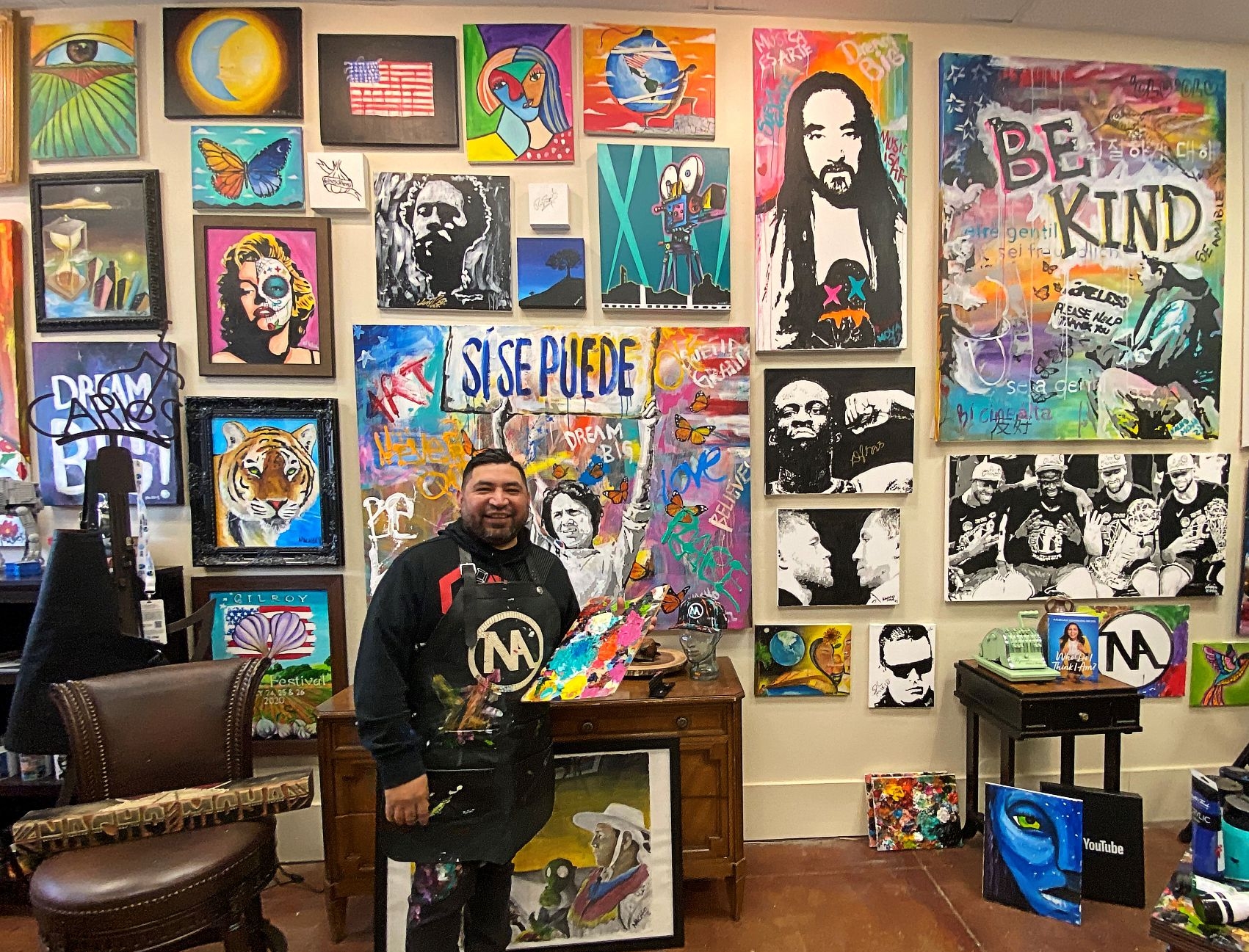 a man holding an artist palate with colorful paintings on the wall behind him