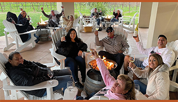 Group of people around a pit fire smiling and holding wine glasses