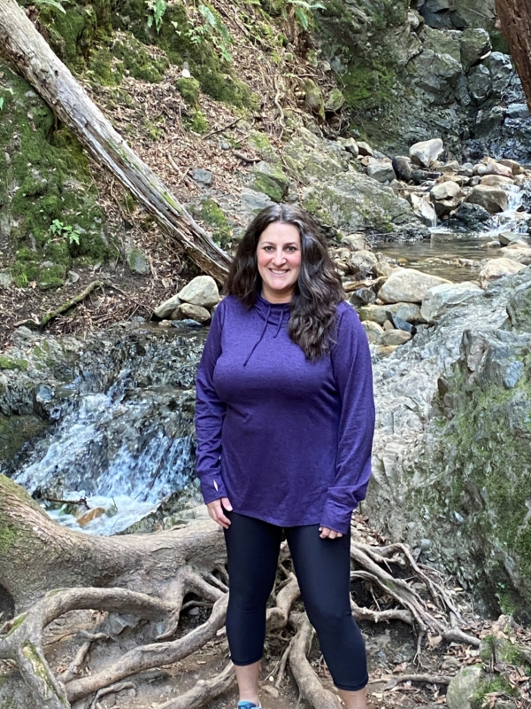 a woman in a purple sweatshirt and black pants posing in front of a waterfall and trees