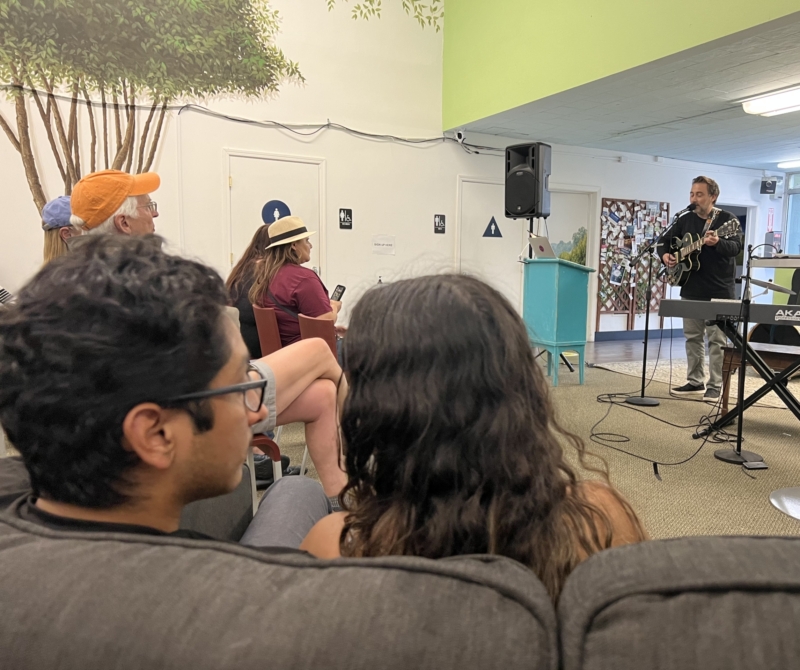 a group of people sit and listen to a man sing and play guitar at a microphone in an indoor space