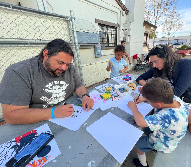 a man, woman, and 2 kids draw while seated at a table outside an art studio