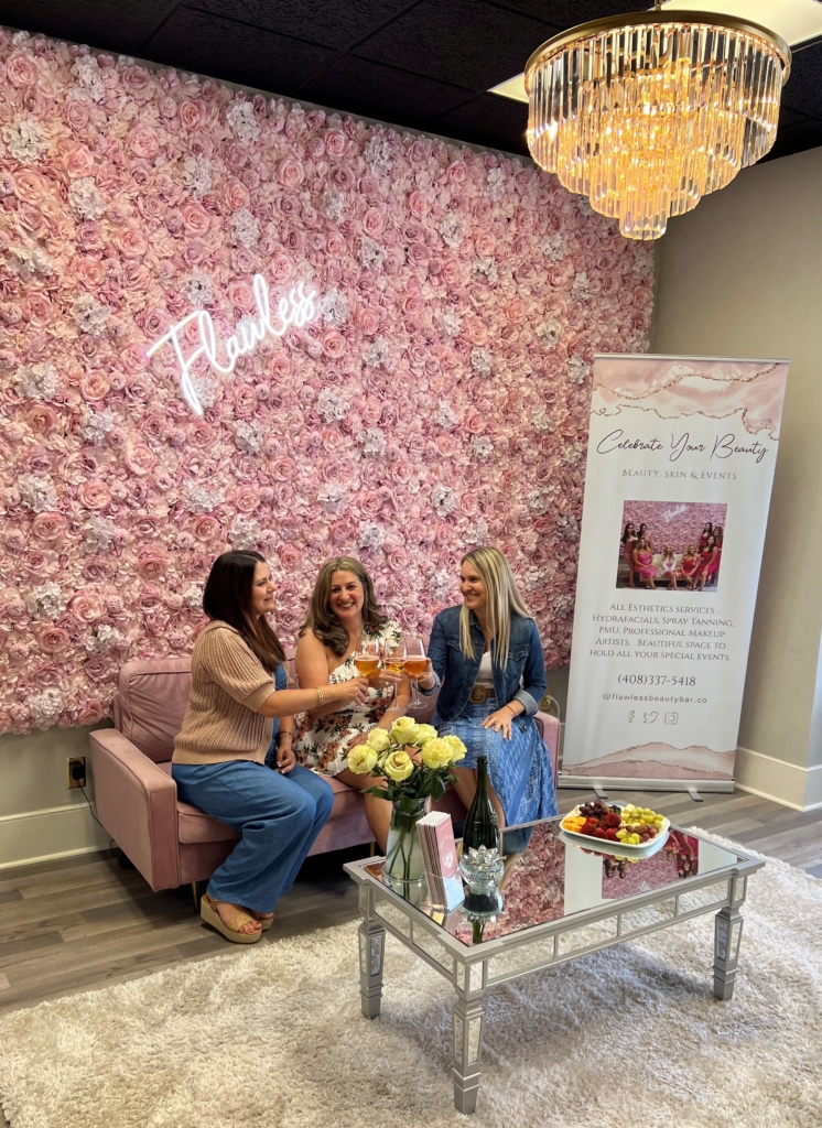 three ladies holding champagne sit on a pink sofa with a pink floral backdrop, a white carpet and silver mirrored table in front of them, and a fancy chandelier above