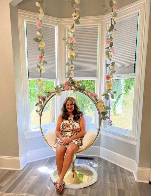 a lady sits in a circular chair with flowers hanging overhead