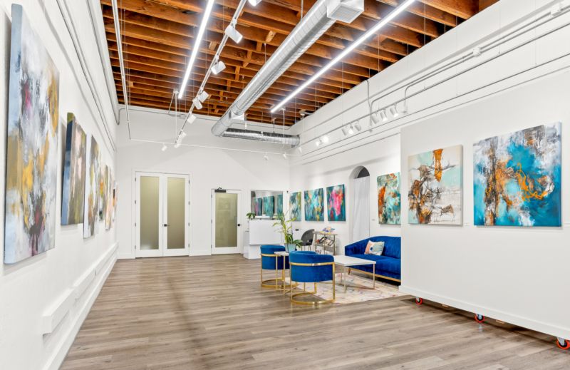 a gallery with white walls and blue seating has several colorful abstract paintings on the walls