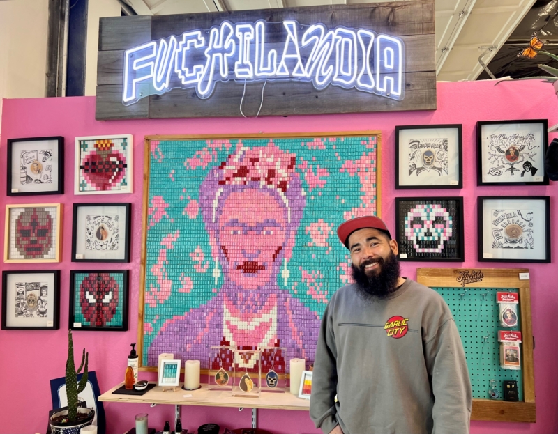 an artist smiles next to his studio sign and his colorful art displayed on a pink wall