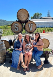 three ladies smile sitting on a bench with wine barrels behind them and grapevines