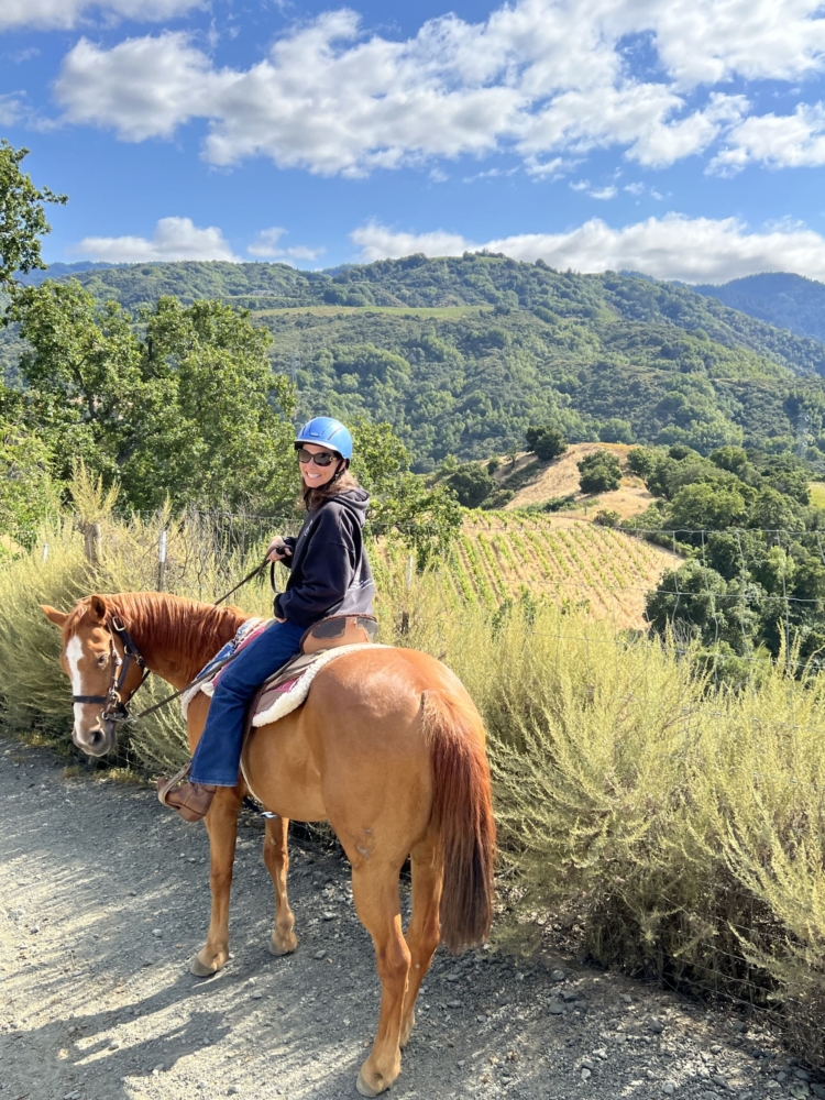 a woman on a brown horse smiles with tree-covered hills and a vineyard in the background