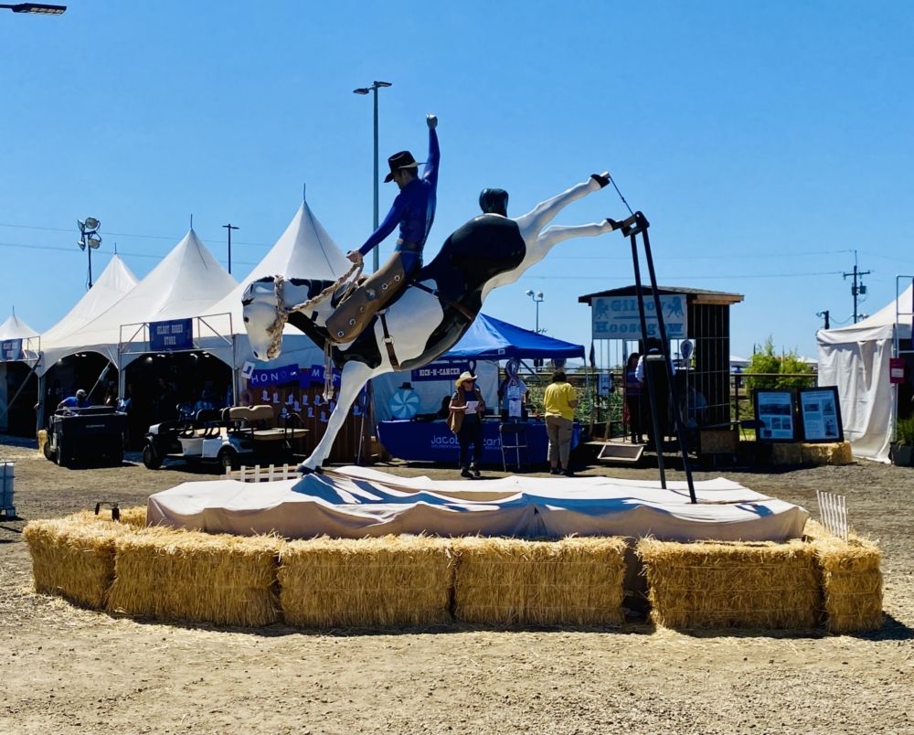 a plastic life-size model of a rodeo cowboy on a bucking horse surrounded by hay bales and vendor tents in the background