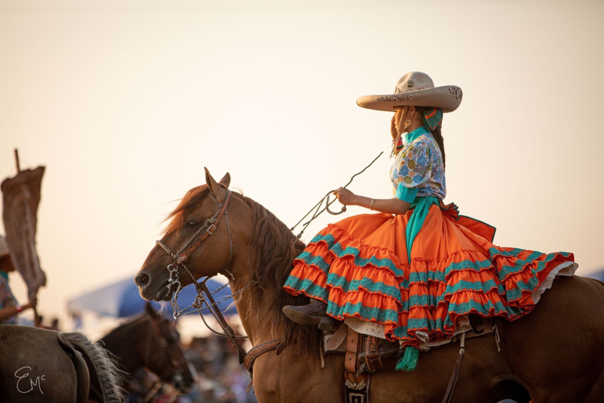 a young girl sits side saddle on a horse in a traditional Mexican dress of orange and green