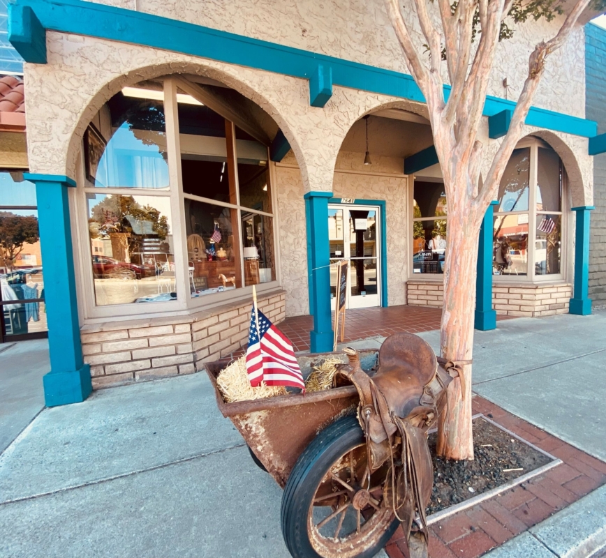 a wheelbarrow with hay and a saddle and American flag sits on the sidewalk outside a beige and turquoise store