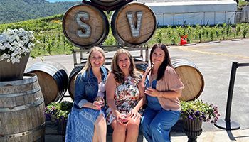 three ladies smile sitting on a bench with wine barrels behind them and grapevines
