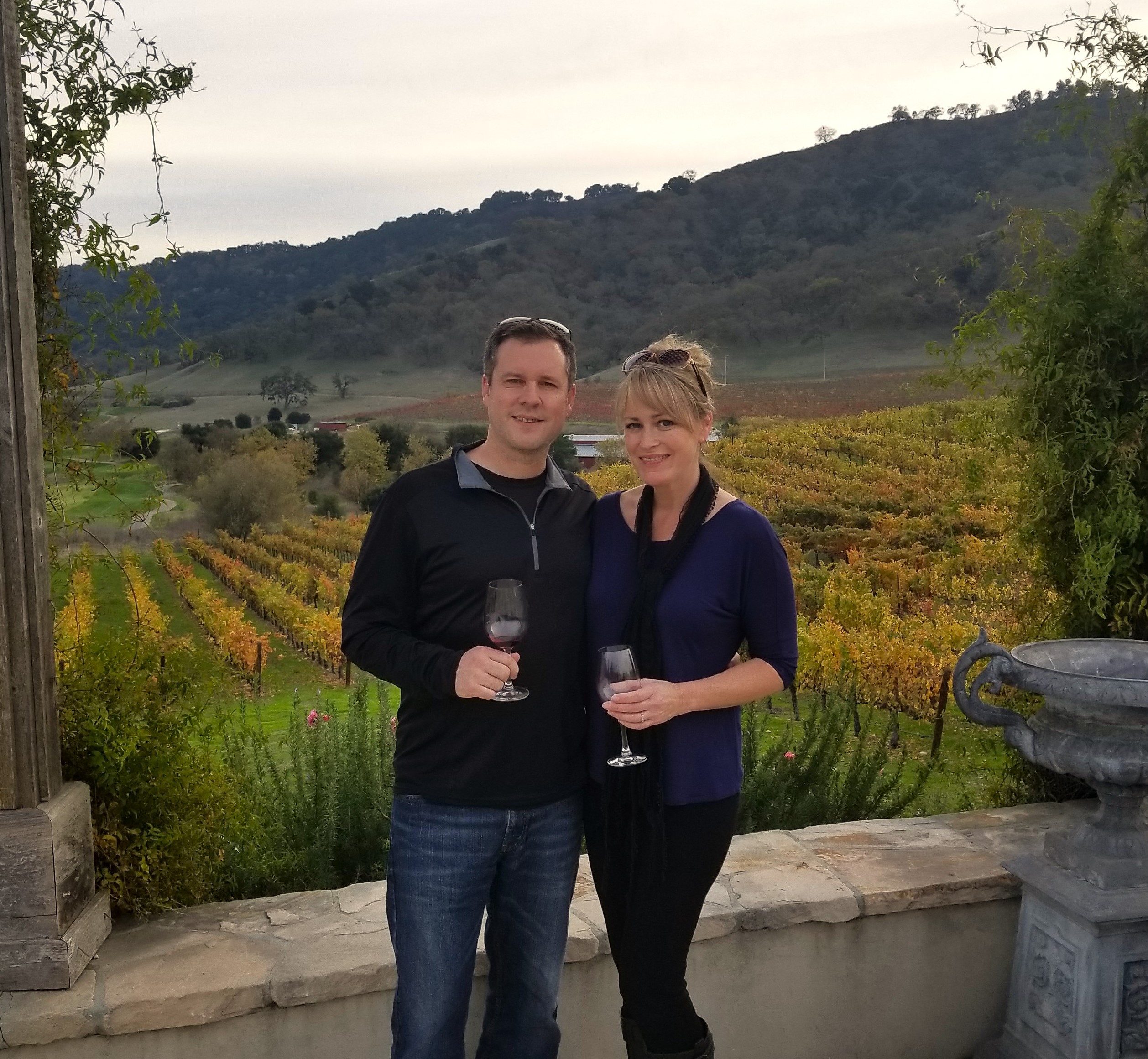 Couple standing together with wine glasses in front of vineyard scenery during harvest season.