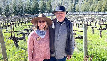 Man and women smiling while standing in front of a green vineyard